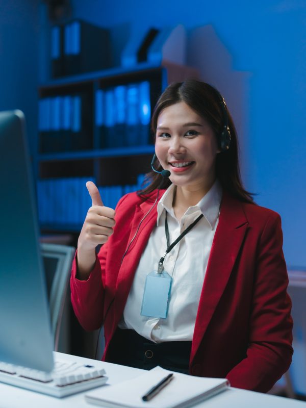 Young asian customer service representative working late at night in office showing thumbs up