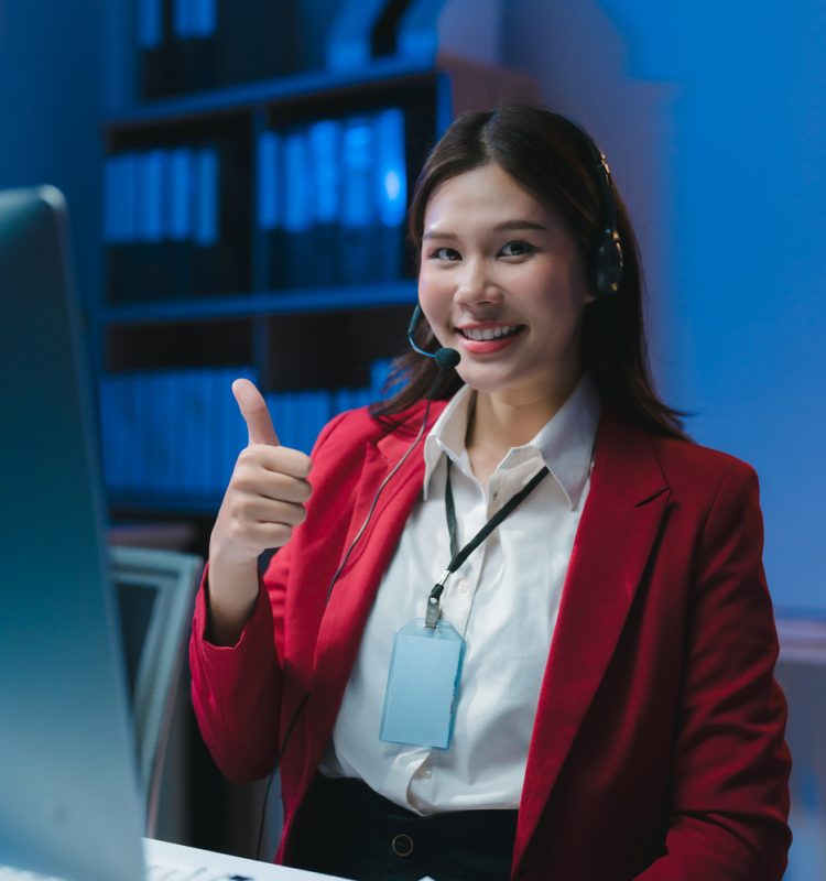Young asian customer service representative working late at night in office showing thumbs up