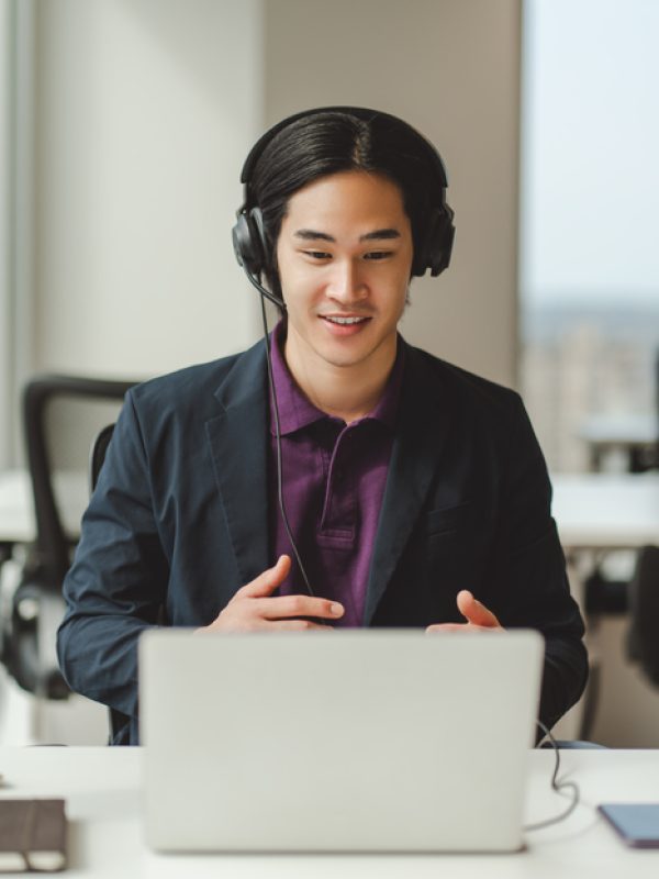 Portrait of confident Asian businessman, call center operator in headset, using laptop, having video call. Attractive Japanese manager working in modern office. Business concept