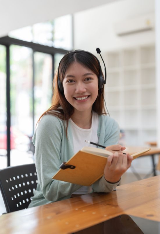 Portrait young asian woman with headphone writing making list taking notes in notepad working or learning on laptop indoors- educational course or training, seminar, education online concept.