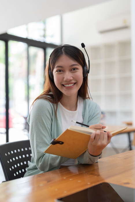 Portrait young asian woman with headphone writing making list taking notes in notepad working or learning on laptop indoors- educational course or training, seminar, education online concept.